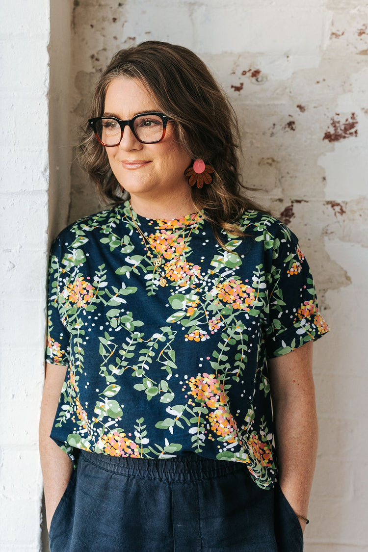 White woman with long dark hair and glasses is standing in front of a white wall wearing a floral top